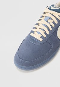 Baskets en suède bleu avec un laçage crème, des détails surpiqués et un logo reconnaissable sur une languette bleu foncé.