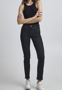 Jeans de mezclilla oscuros de talle alto y corte ajustado, con cinco bolsillos, costuras en contraste y cierre de botón. Combinados con una camiseta sin mangas negra y zapatillas blancas.