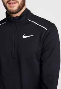 Czarna sportowa bluzka z zamkiem Nike z wysokim kołnierzem, z białymi akcentami w postaci pasków oraz małym białym logo na piersi. Teksturowana tkanina.