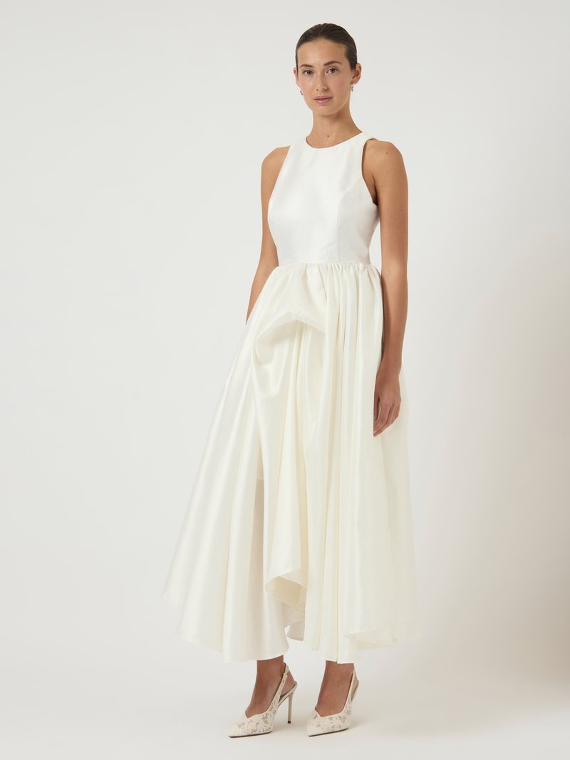 Γυναίκα με λευκή σατέν μίντι φόρμα χωρίς μανίκια και φούστα με πτυχώσεις, φορώντας φλοράλ δαντελένια ψηλοτάκουνα σαμπό, που στέκεται σε άσπρο φόντο.