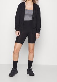 Sudadera negra con cremallera sobre una camiseta sin mangas gris, combinada con pantalones cortos negros y zapatillas negras con suelas texturizadas. Unas calcetas negras completan el look.