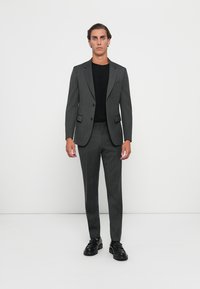 Grijze pak in een getailleerde pasvorm met een notch lapel, bijpassende broek en een zwart shirt. Heeft subtiele krijtstrepen en een standaard knoopsluiting.