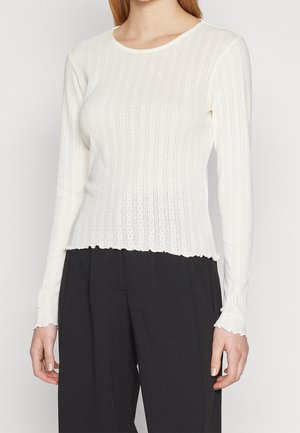 Femme portant un top en maille côtelée blanche à manches longues avec des bords festonnés et un pantalon taille haute noir.