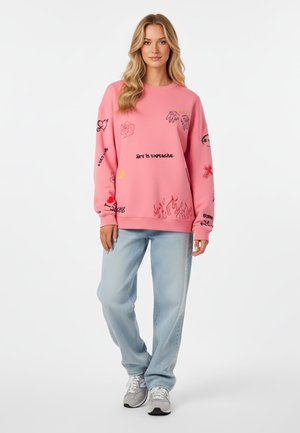 Roze fleece sweatshirt met verschillende grafische prints in zwart en geel, gecombineerd met lichtblauwe, loszittende jeans en grijze sportschoenen.