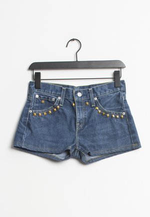 Shorts en denim de couleur bleue, avec des accents de clous dorés sur les poches avant et des coutures jaunes contrastantes. Accrochés à un cintre noir.
