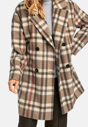 Frau trägt einen knielangen, doppelt geknöpften karrierten Mantel in Beige, Braun und Weiß mit schwarzen Knöpfen über einem grauen Rollkragenpullover.