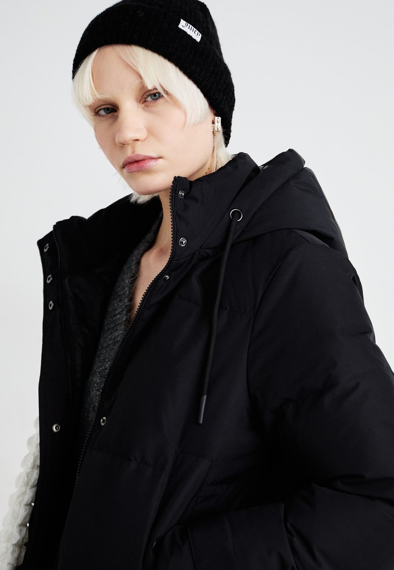 Schwarze Pufferjacke mit Kapuze, ausgestattet mit Reißverschlüssen und Knöpfen, über einem grauen Pullover. Das Modell trägt eine gestrickte schwarze Mütze.