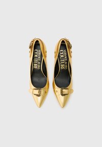 Gold pointed-toe shoes feature a shiny leather material, decorative buckle accents, and a black insole with logo detailing.