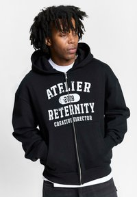 Crna dukserica na patentni zatvarač, izrađena od mješavine pamuka, s prednjim printom "ATELIER RETERNITY 2018 KREATIVNI DIREKTOR" u bijeloj boji, rebrastim rukavima i džepovima.