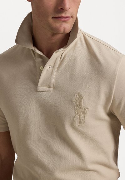 Beige polo paita valmistettu teksturoidusta kankaasta, jossa on kaulus ja kaksi nappia. Vasemmassa rinnassa brodeerattu polo-pelaajan logo.