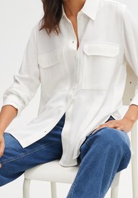 Femme assise portant une chemise blanche ample à boutons avec deux poches poitrine et manches retroussées, associée à un jean bleu.