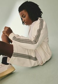 Hvid tekstureret træningsdragt med sorte striber, parret med hvide sneakers og sorte sokker, siddende på en lys overflade.