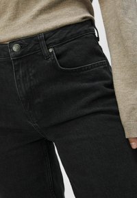 Jeans de mezclilla negros con un ajuste slim, que cuentan con un bolsillo frontal, cierre de botón y una textura sutil. Combinados con un suéter beige claro.
