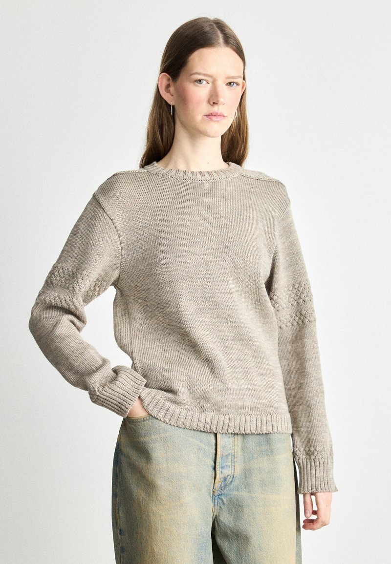 Beige knitted sweater with round neckline, textured accents on sleeves, and ribbed cuffs and hem, paired with light blue denim jeans.