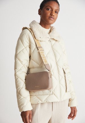 Person iført en cremefarvet quiltet jakke med en lammeskindskrave og beige bukser, bærende en taupe crossbody taske med en bred rem.