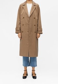 Persona vestida con un abrigo de trench largo color tostado y de doble botonadura, camiseta blanca, jeans azules cortos y tacones negros con correa al tobillo, contra un fondo blanco.