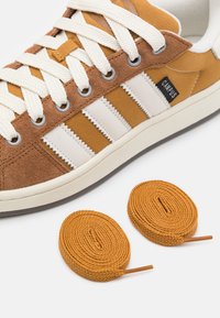Sneaker marrone e beige con strisce bianche, realizzato in suede e tela. Include due lacci arancioni rotondi intrecciati con punte metalliche.