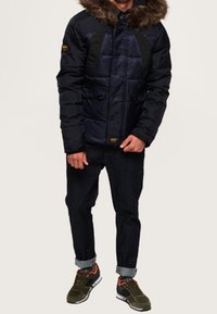 Giacca a vento blu scuro con dettagli neri e cappuccio foderato in pelliccia, dotata di tasche frontali e chiusura con bottoni, indossata sopra jeans scuri e sneakers.