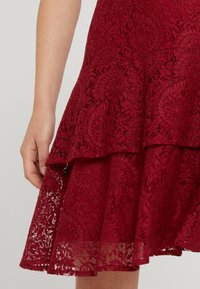 Main reposant à côté d'une jupe bordeaux en dentelle, longueur genou, avec un motif floral et des volants superposés.