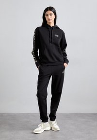 Ensemble de hoodie noir et de survêtement en tissu doux. Présente des accents de logo dorés sur la poitrine et le long des manches. Baskets blanches portées.