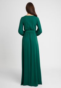 Długa, zwiewna sukienka w jednolitym, szmaragdowozielonym kolorze. Posiada długie rękawy, dopasowaną talię z paskiem oraz kwadratowy dekolt. Gładka faktura.