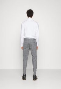 Uomo con capelli ricci che indossa una camicia bianca a maniche lunghe, pantaloni a quadri grigi e stivali neri, in piedi di fronte a un muro bianco semplice.
