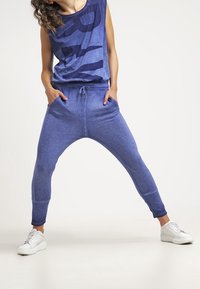 Blå ärmlös tröja med grafisk design, parat med lösa blå joggers. Båda plaggen har en mjuk textur, kompletterat med vita sneakers.