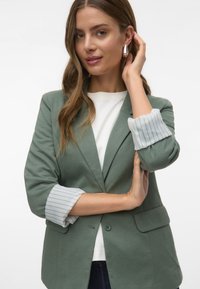 Groene blazer met een gestructureerd ontwerp, voorzien van gestreepte witte en groene voering aan de manchetten. Lichtgewicht stof en twee knopen sluiting.