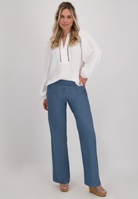 Witte blouse van gestructureerde stof met haakdetails bij de halslijn, gecombineerd met blauwe wijde broek en tan gekleurde sleehaksandalen.