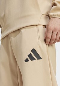 Beige sporta bikses no teksturēta auduma, ar lielu melnu Adidas logo kreisajā augšstilbā un roku kabatā.