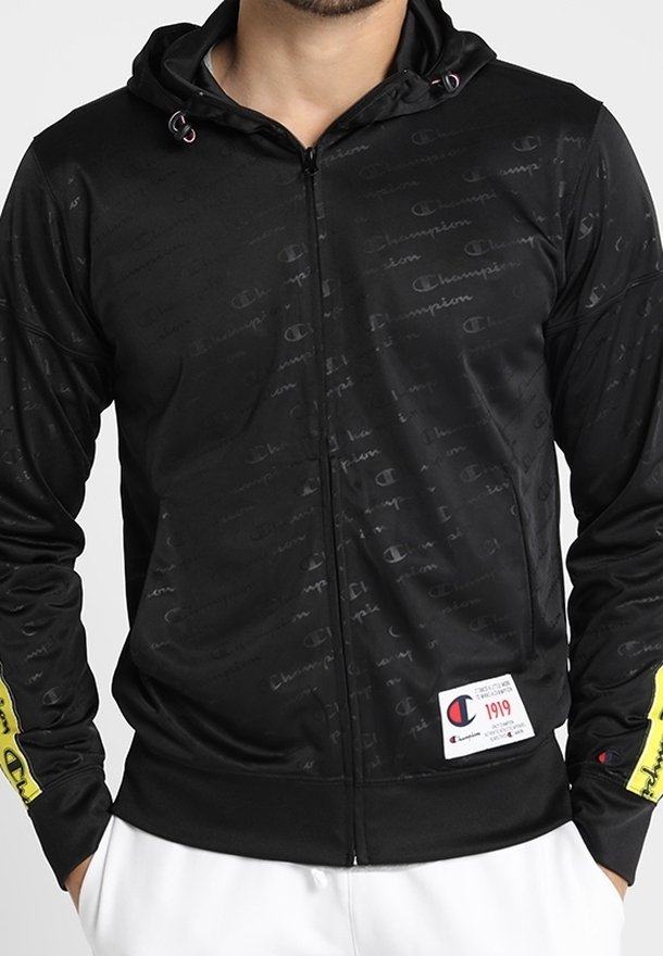 Svart zip-up huva jacka med texturerad tyg, med subtil logotypmönster, gula detaljer på ärmarna och en liten etikett i nederkant.