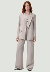 Hellgrauer Anzug mit maßgeschneiderter Passform, bestehend aus einem langen Blazer mit Reverskragen, einer knopfbaren Weste und weit geschnittenen Hosen. Glatter Stoff mit minimalen Akzenten.