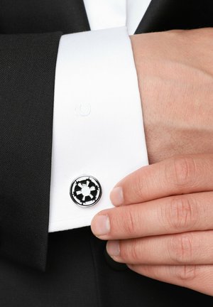 Spinki do mankietów na białej koszuli z czarnym garniturem. Spinki posiadają okrągły wzór z chromowym wykończeniem i czarnymi akcentami.