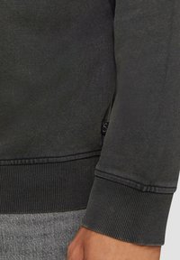 Czarna bluza z ribbami przy mankietach i hemie, o miękkiej teksturze, z widoczną metką po boku. Materiał wydaje się casualowy i wygodny.