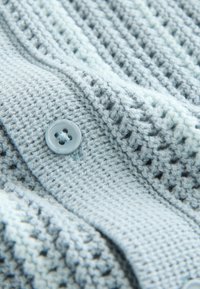 Light blue knitted fabric with a close-up of a round button sewn onto ribbed and textured knit patterns.