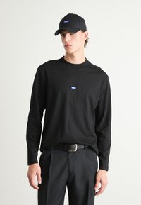 Zwarte langewijl t-shirt met een klein blauw logo op de borst, gecombineerd met zwarte broek en een bijpassende zwarte pet. Eenvoudig en hedendaags ontwerp.