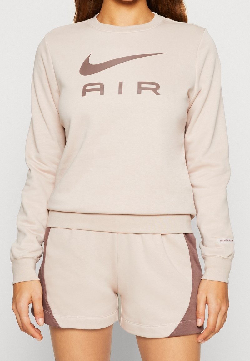 Jasnobeżowa bluza z kapturem z brązowym logo Nike, zestawiona z beżowymi szortami z brązowymi bocznymi akcentami. Miękki materiał o swobodnym kroju.