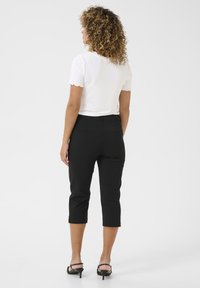 Vrouw met krullend haar draagt een witte top met korte mouwen, zwarte cropped broek en zwarte hoge hakken, staand tegen een effen achtergrond.