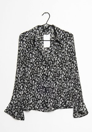 Schwarz-weiße, abstrakt gemusterte Bluse mit langen Ärmeln, Knöpfen, Kragen und ausgestellten Manschetten, die an einem schwarzen Kleiderhanger vor einem weißen Hintergrund hängt.