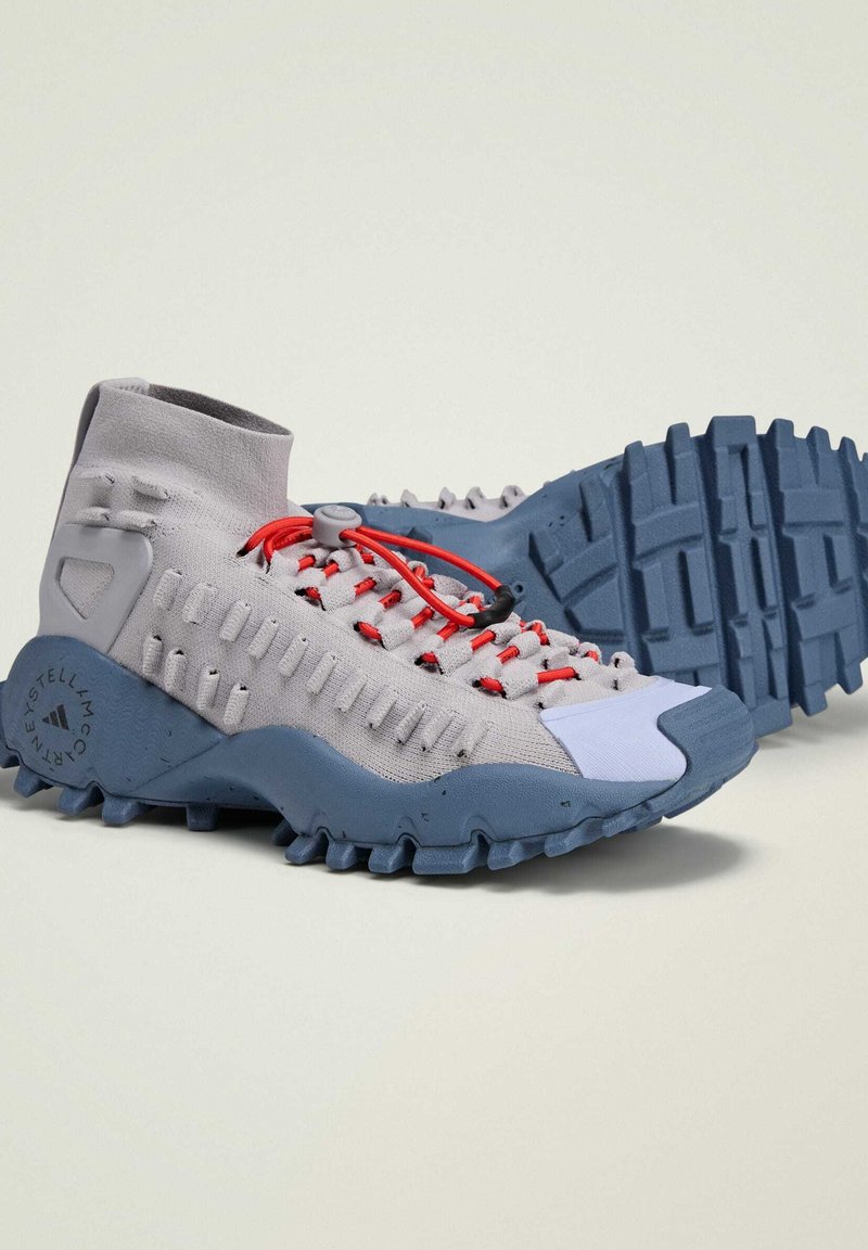 Scarpe alte grigie con una superficie testurizzata, lacci elastici rossi e una suola blu robusta con battistrada prominenti e un dettaglio del logo.