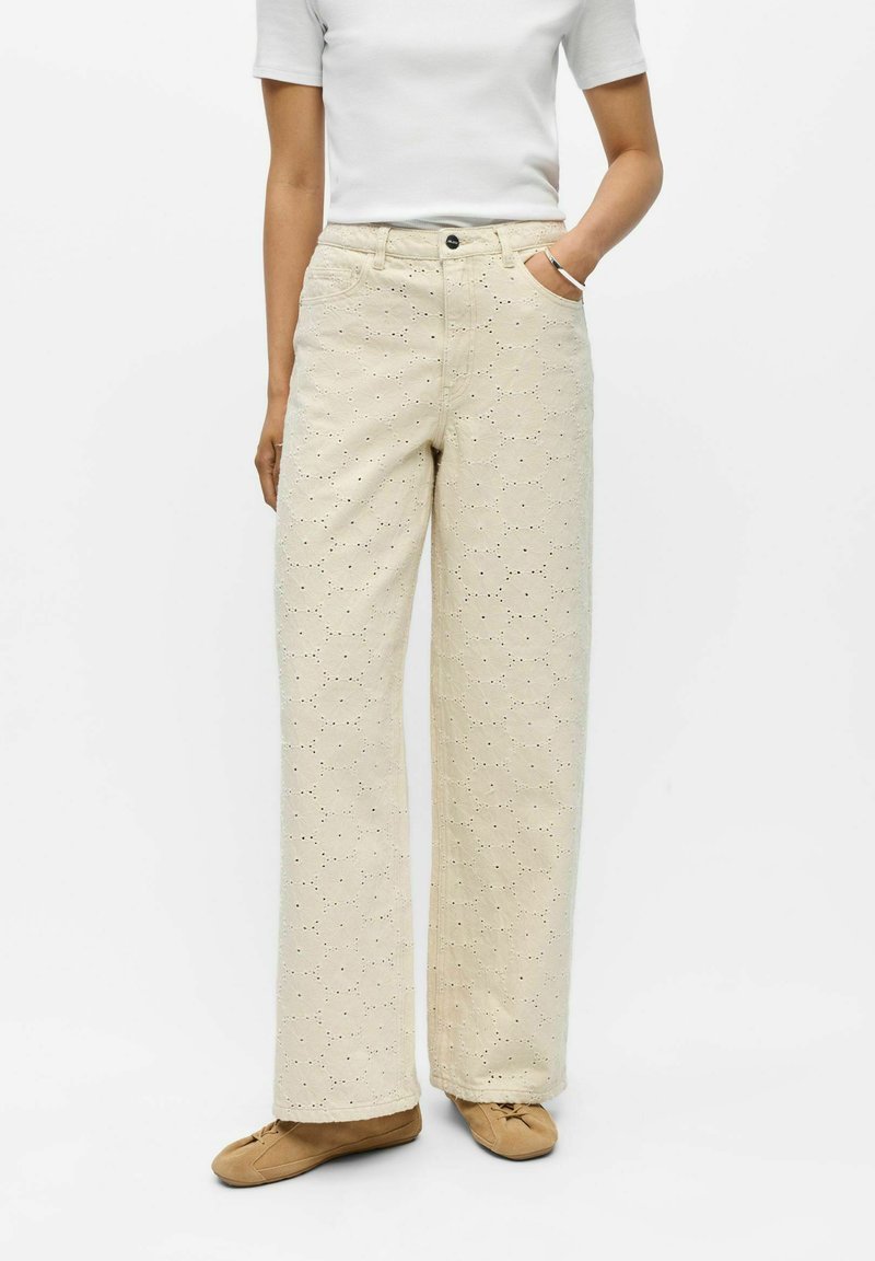 Femme portant un pantalon beige à jambes larges avec broderie ajourée, un haut blanc à manches courtes et des chaussures plates couleur camel, debout avec une main dans la poche.