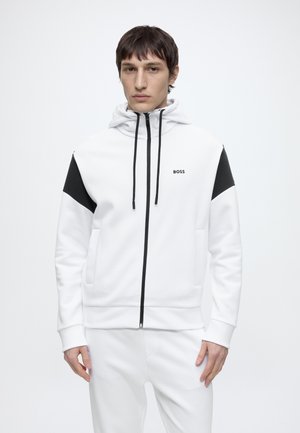 Man die een witte hoodie met ritssluiting draagt met zwarte accenten op de schouders en zwarte trekkoorden, staand tegen een effen witte achtergrond.