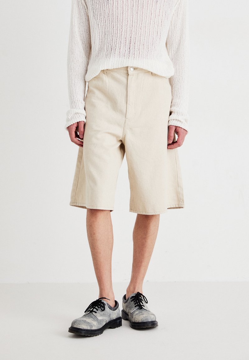 Shorts en denim beige à coupe décontractée, avec un devant plat et des poches latérales cousues, associés à un pull en tricot blanc texturé et des chaussures grises.