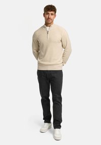 Beige strikket pullover med en halv lynlås krave, ribbede manchetter og kant, parret med mørke grå jeans og lyse sneakers på en ensfarvet baggrund.
