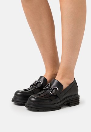 Mocassins en cuir noir avec détail mors en métal argenté et franges, portés pieds nus sur un fond blanc uni.