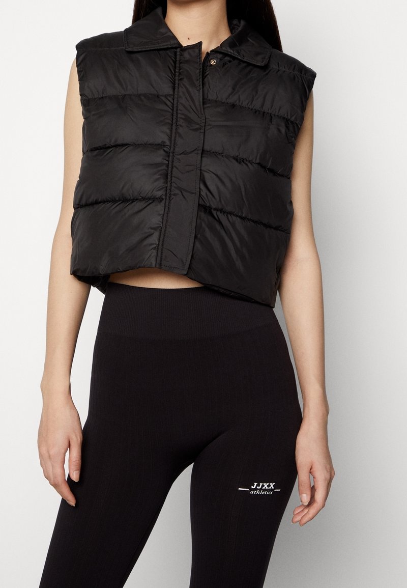 Schwarze, kurz geschnittene Steppweste mit horizontalen Steppnähten, einem Kragen und einem Druckknopfverschluss. Trägt man über schwarzen, gerippten Leggings mit Logodetail.