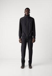 Black athletic set featuring a zippered jacket with a high collar and elastic cuffs, paired with tapered pants. Smooth, lightweight fabric.