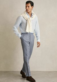 Cămașă de in albastru deschis, pantaloni gri ajustați, pulover crem drapat elegant și pantofi din Suede maro. Textură netedă cu un design clar.