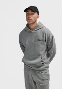 Młody mężczyzna w szarej bluzie z kapturem i pasujących dresach z czarnym napisem "HALO 196819721981", oraz czarnej czapce, stojący na białym tle.