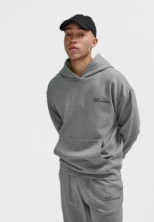 Junger Mann, der einen grauen Hoodie und passende Jogginghosen mit schwarzem Text "HALO 196819721981" sowie eine schwarze Mütze trägt, vor einem weißen Hintergrund steht.